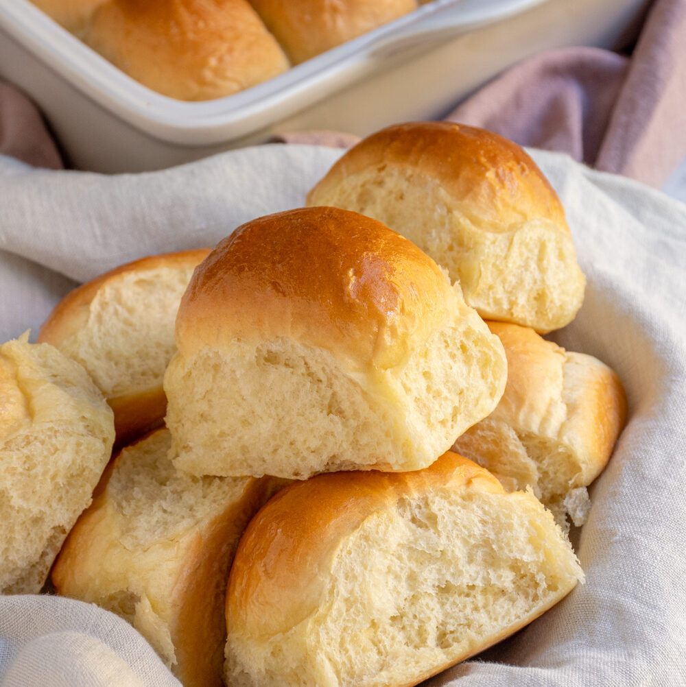 A linen lined basket filled with a bunch of golden brown soft dinner rolls.
