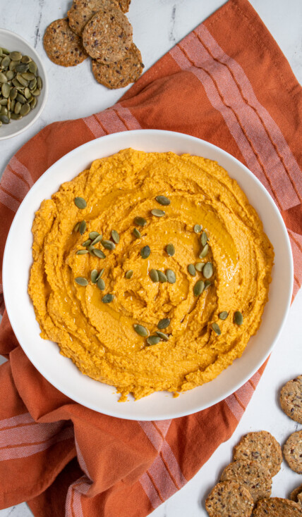 Overhead view of a large white bowl filled with pumpkin hummus and garnished with olive oil and pumpkin seeds.
