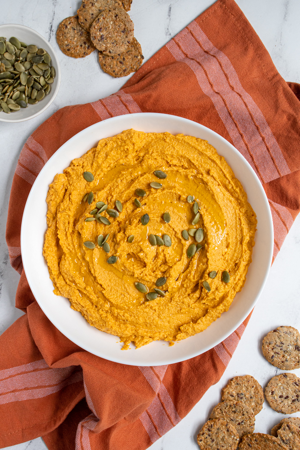 Overhead view of a large white bowl filled with pumpkin hummus and garnished with olive oil and pumpkin seeds.