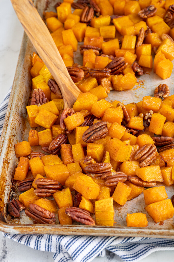 Sheet pan with roasted maple butternut squash with pecans, some scooped together with a wooden spatula.