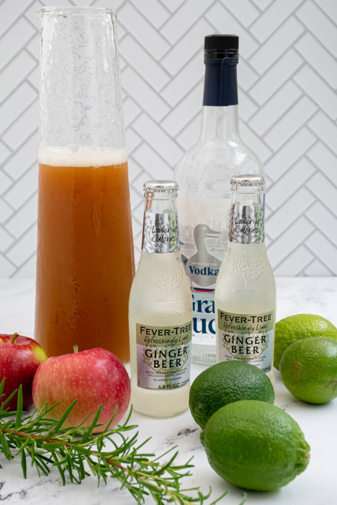 Ingredients needed to make apple cider Moscow mules laid out on a table including limes, apples, ginger beer, apple cider, and vodka.