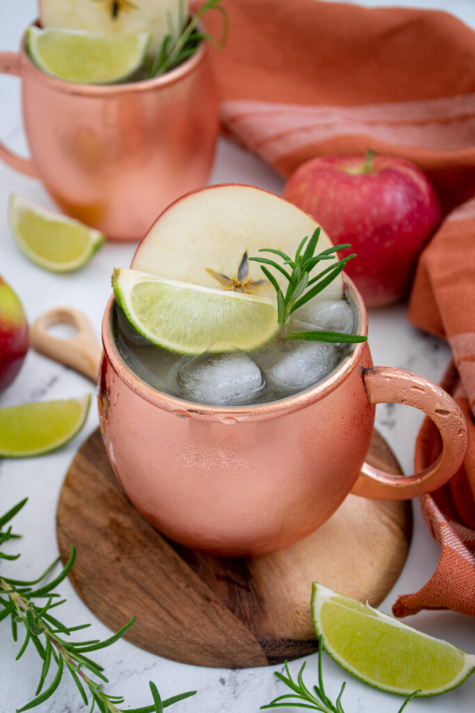 Copper mugs with apple cider mules garnished with an apple wheel, lime wedge, and rosemary sprig.