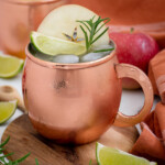 Pinterest pin with logo overlay 'The Schmidty Wife', image of a copper mug full of apple cider mule garnished with apple, rosemary, and lime.