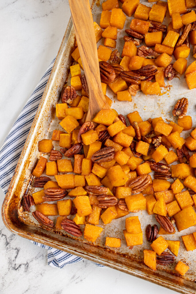 A baking pan filled with diced and cooked butternut squash with pecans , some scooped with a wood spatula.