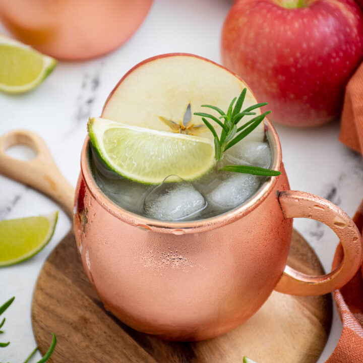 Copper mug on a cutting board filled with apple cider mule and garnished with an apple wheel, lime wedge, and rosemary spring.