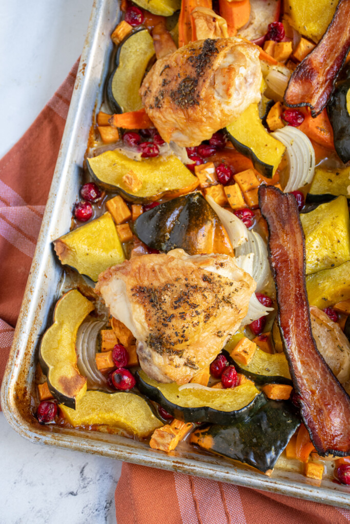 Corner view of a sheet pan with a chicken thigh and other autumn seasonal veggies underneath cooked into a one pan dinner.