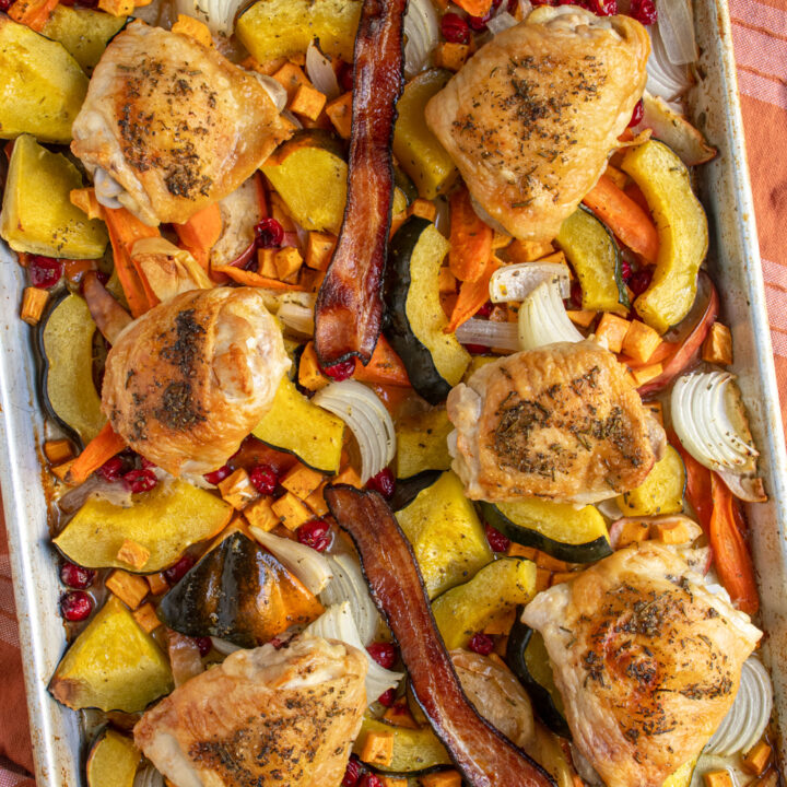 A sheet pan with an autumn chicken dinner including squash, cranberries, onions, chicken thighs and bacon.