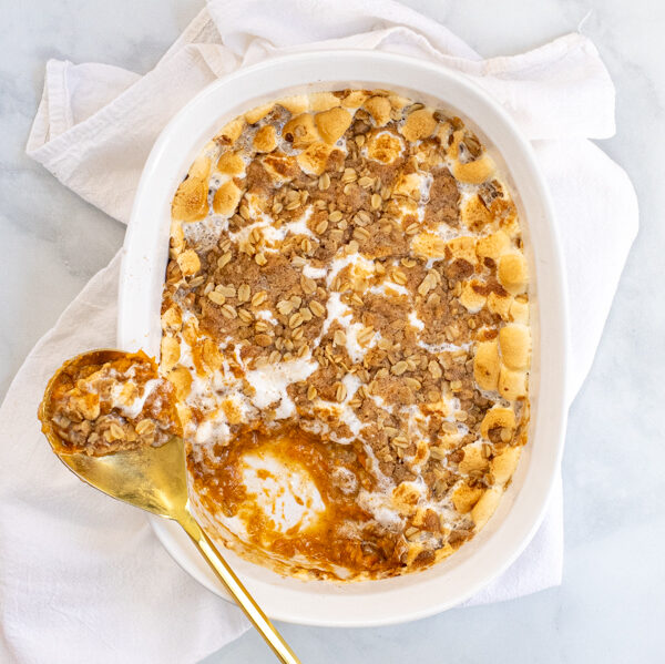 Sweet Potato Casserole with Marshmallows and Oat Streusel in a white baking dish with a gold serving spoon sitting on a white kitchen towel.