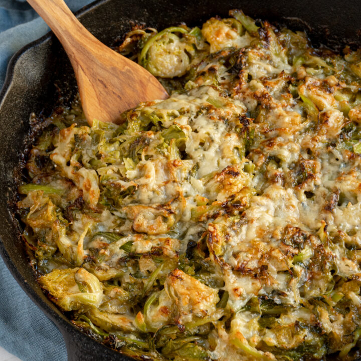 A wooden spoon scooping cheesy brussel sprouts gratin from a skillet.