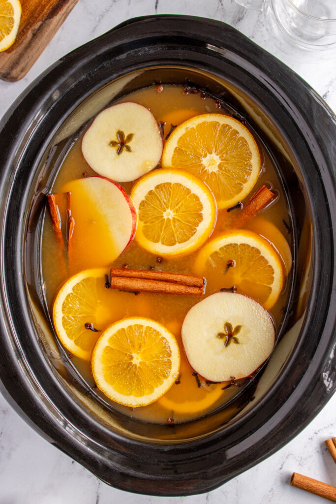 Crockpot filled with Christmas wassail and topped with orange slices, apples slices, cloves, and cinnamon sticks.