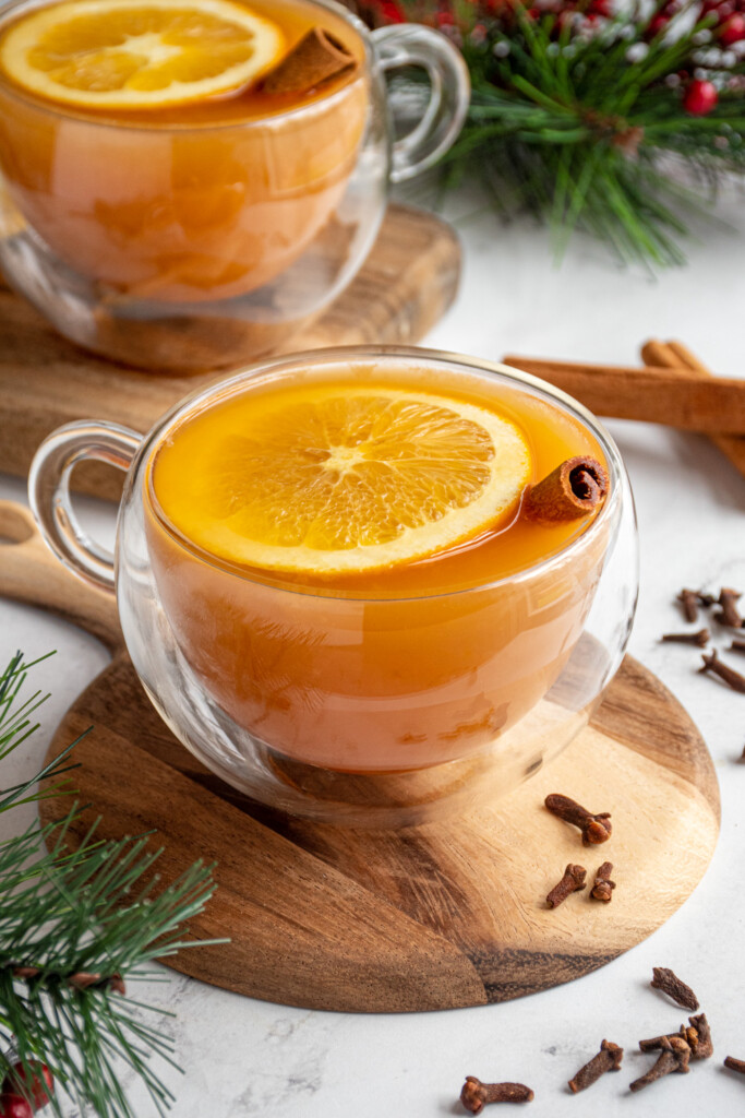 A clear glass mug filled with warm wassail garnished with an orange slice and cinnamon stick with cloves scattered about the table it is on.