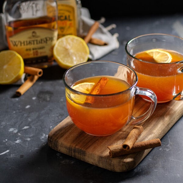 Two clear mugs on a wooden cutting board against a black background filled with hot toddy cocktail and garnished with cinnamon sticks and a lemon wedge.
