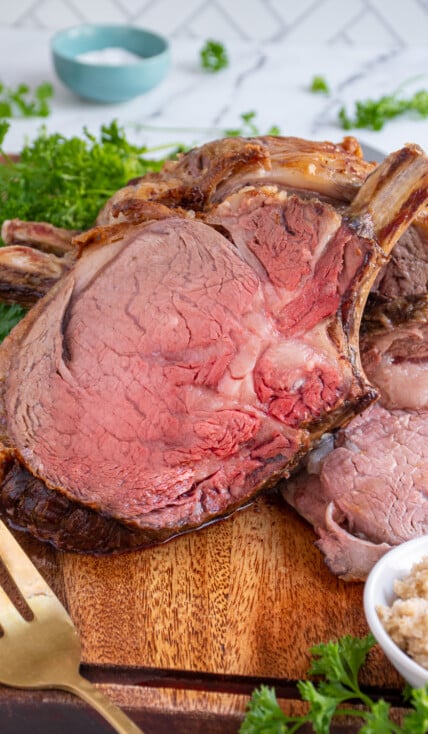 A large slice of prime rib on a cutting board leaned up against the rest of the roast showing off a red tender inside.