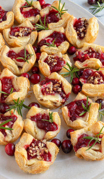 An angled view of a white platter full of cranberry brie bites garnished with finely chopped nuts and sprigs of rosemary.