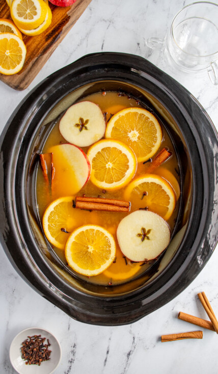 A black crockpot filled with Christmas wassail topped with apple and orange slices, cinnamon sticks, and cloves.