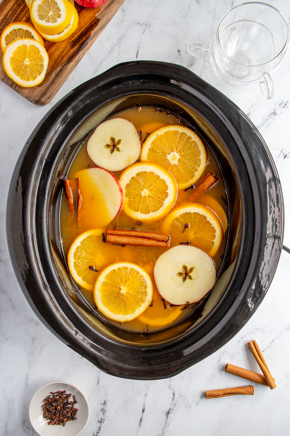 A black crockpot filled with Christmas wassail topped with apple and orange slices, cinnamon sticks, and cloves.