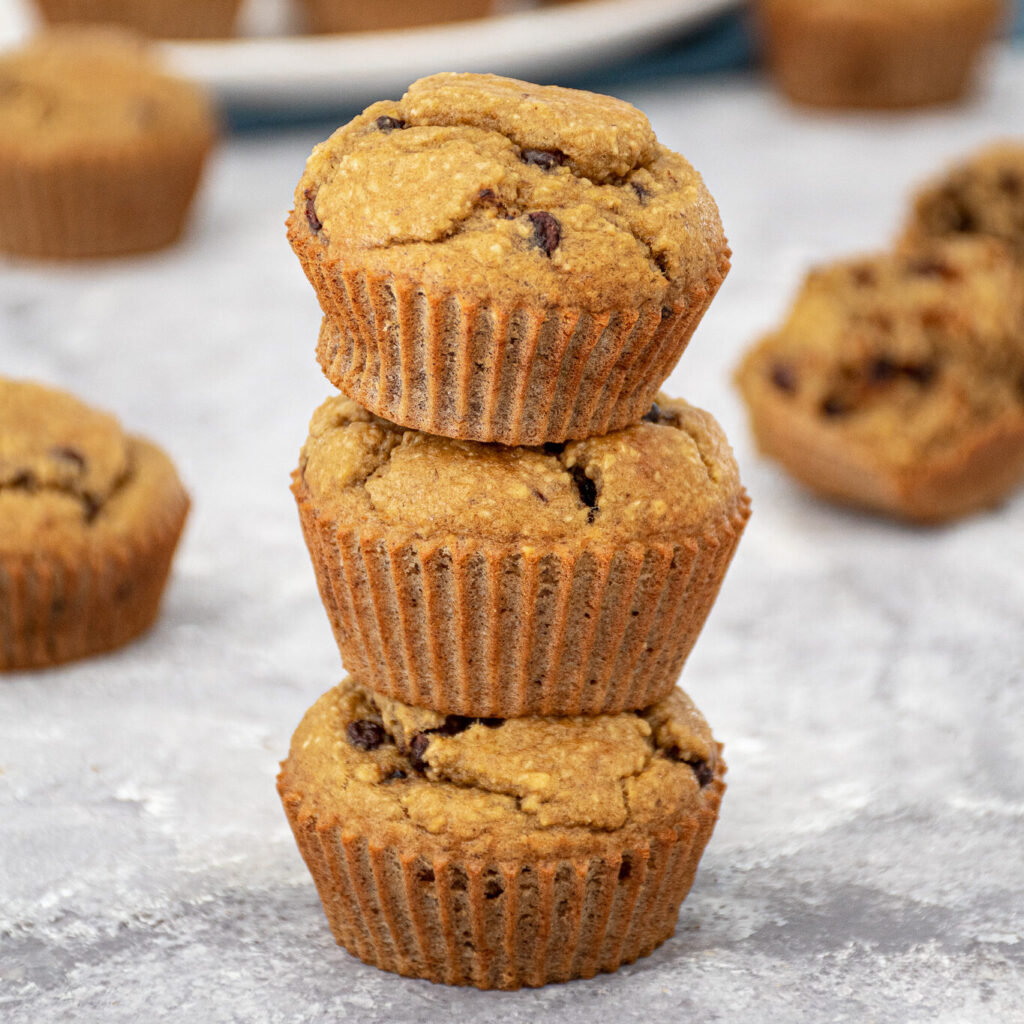 A stack of 3 blender flourless banana muffins with mini chocolate chips making a muffin tower with a plate of muffins in the background.