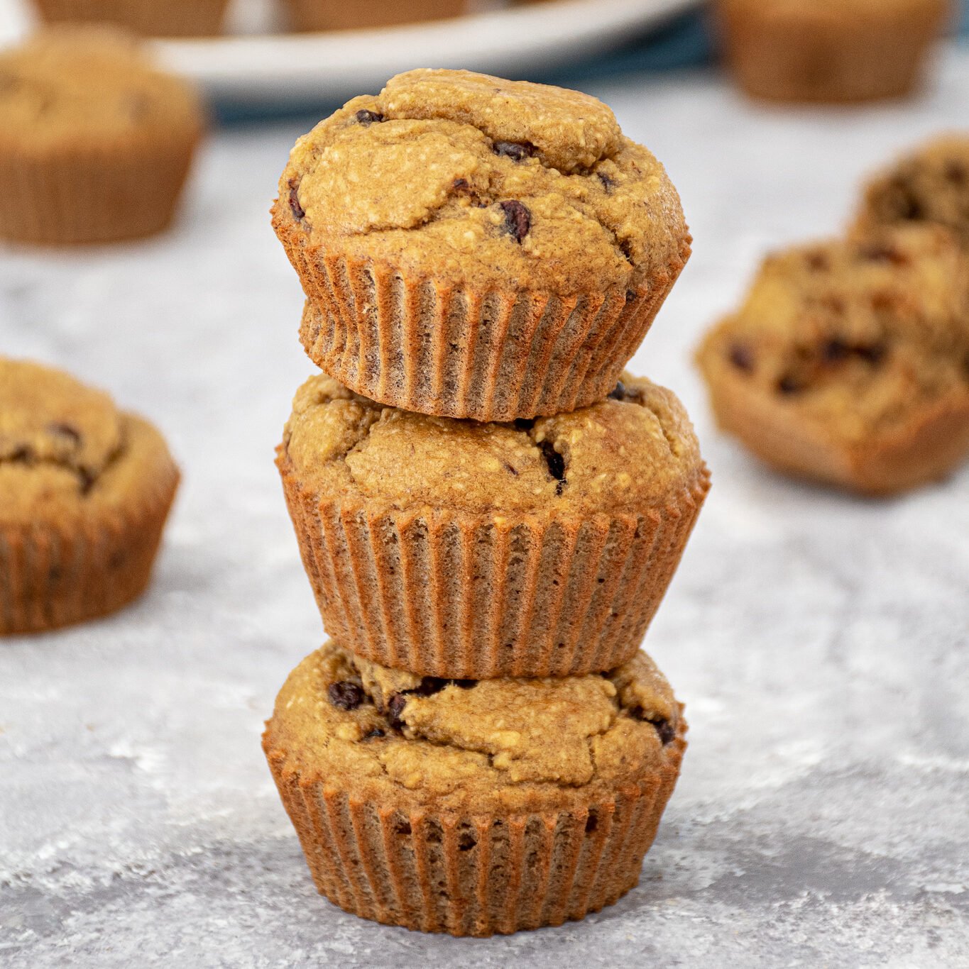 A stack of 3 blender flourless banana muffins with mini chocolate chips making a muffin tower with a plate of muffins in the background.