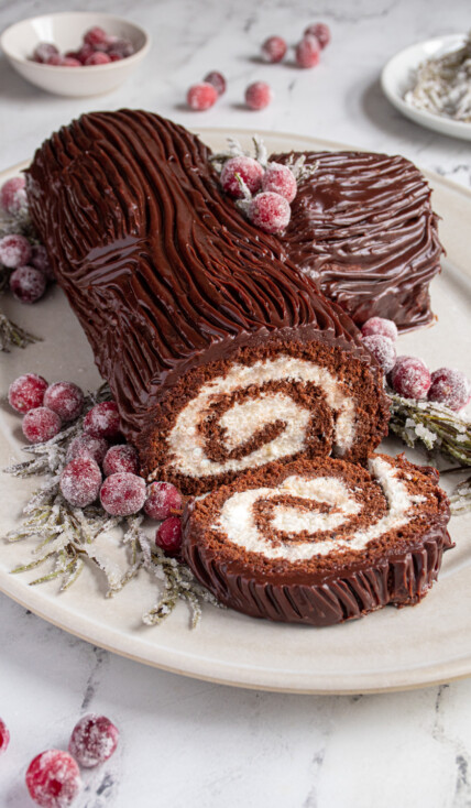Yule Log Cake (Bûche de Noël) laid on a platter with a slice cut off showing the rolled up inside, garnished with sugared cranberries and rosemary.