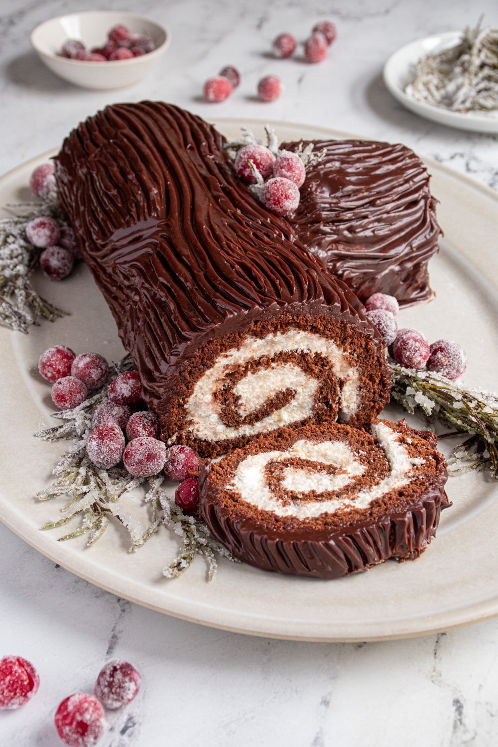 Yule Log Cake (Bûche de Noël) laid on a platter with a slice cut off showing the rolled up inside, garnished with sugared cranberries and rosemary.