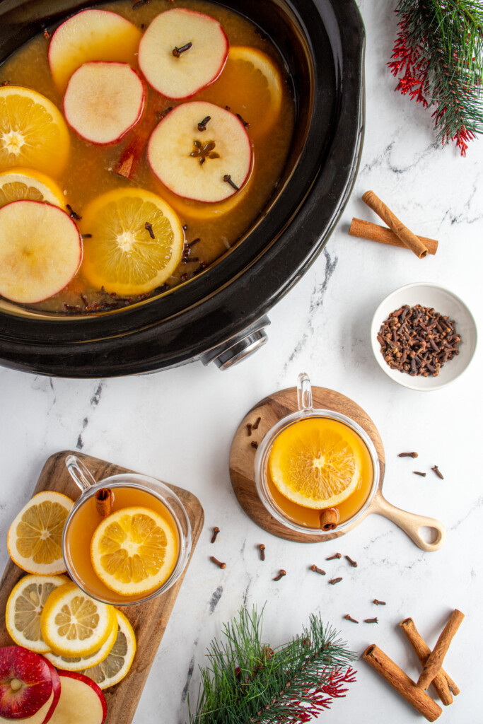 Overhead view of wassail in a crockpot next to full glasses of Christmas wassail in clear mugs topped with orange slices and cinnamon sticks.