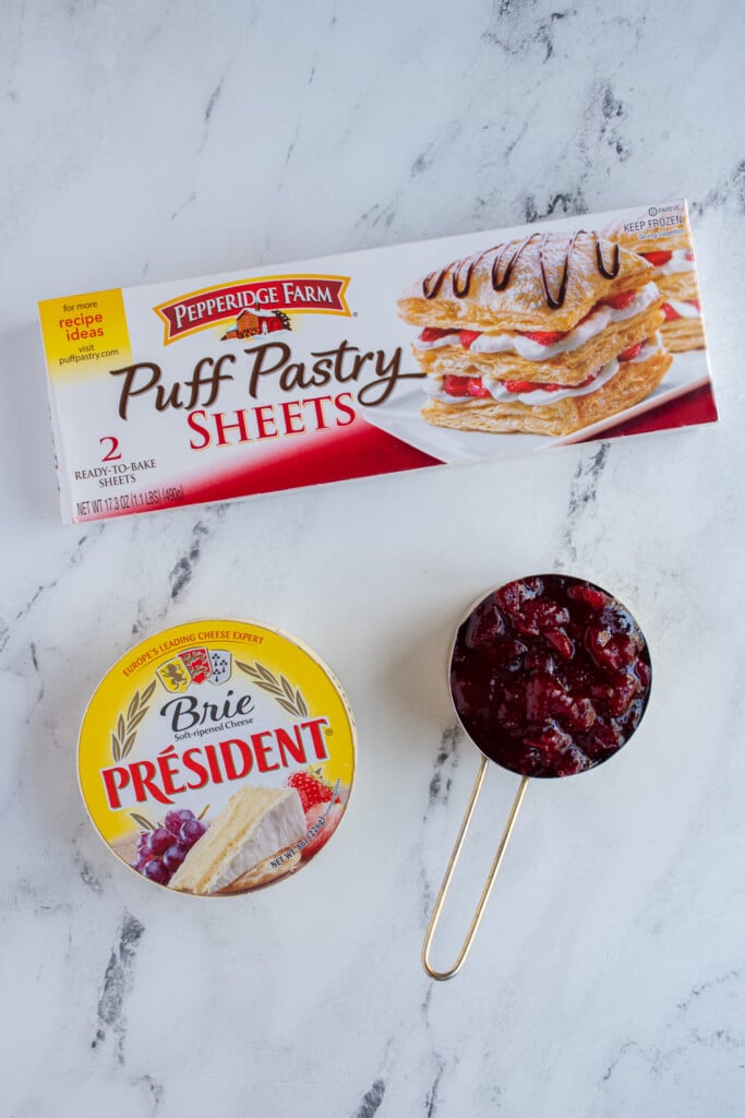 Ingredients needed to make cranberry brie bites on a tabletop including puff pastry, brie cheese, and cranberry sauce.