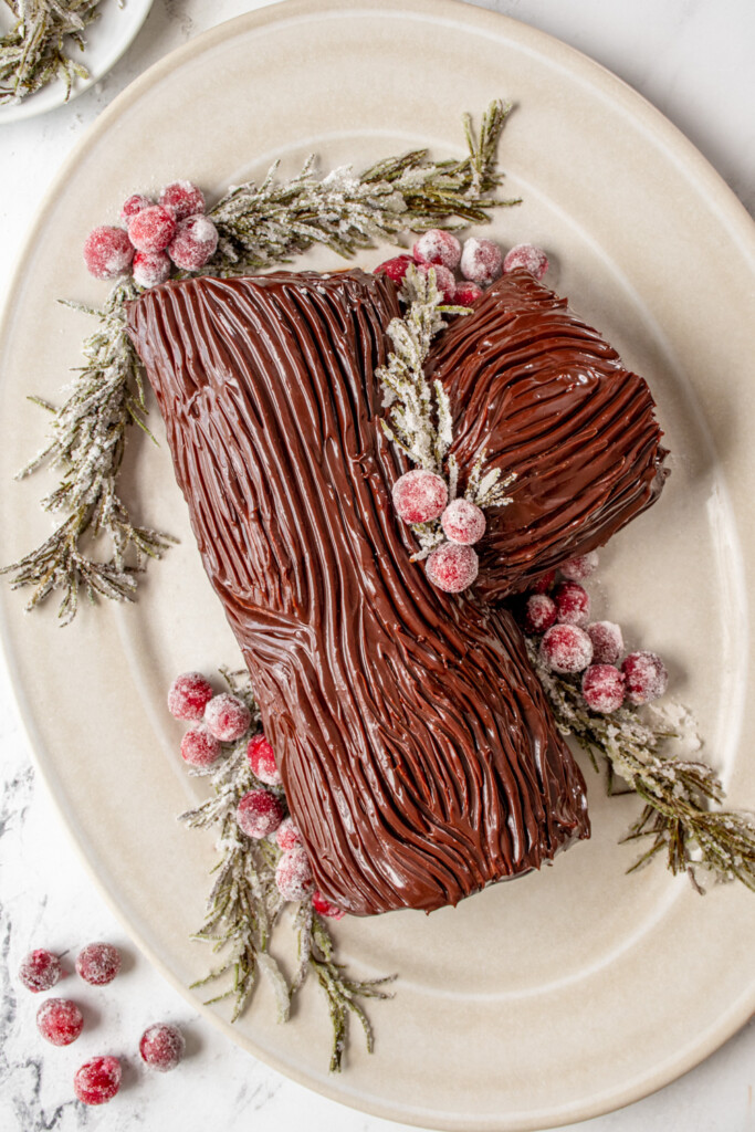 A yule log cake on a white platter with chocolate ganache that looks like tree bark and garnished with cranberries and rosemary covered in sugar.