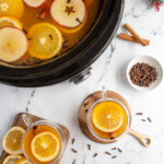 Pinterest Pin with logo overlay 'The Schmidty Wife', image of a crockpot and mugs filled with wassail and garnished with orange slice and spices.