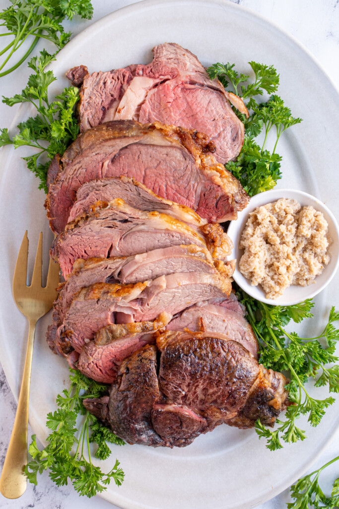 White platter full of cut prime rib ready to serve along with horseradish and fresh parsley.