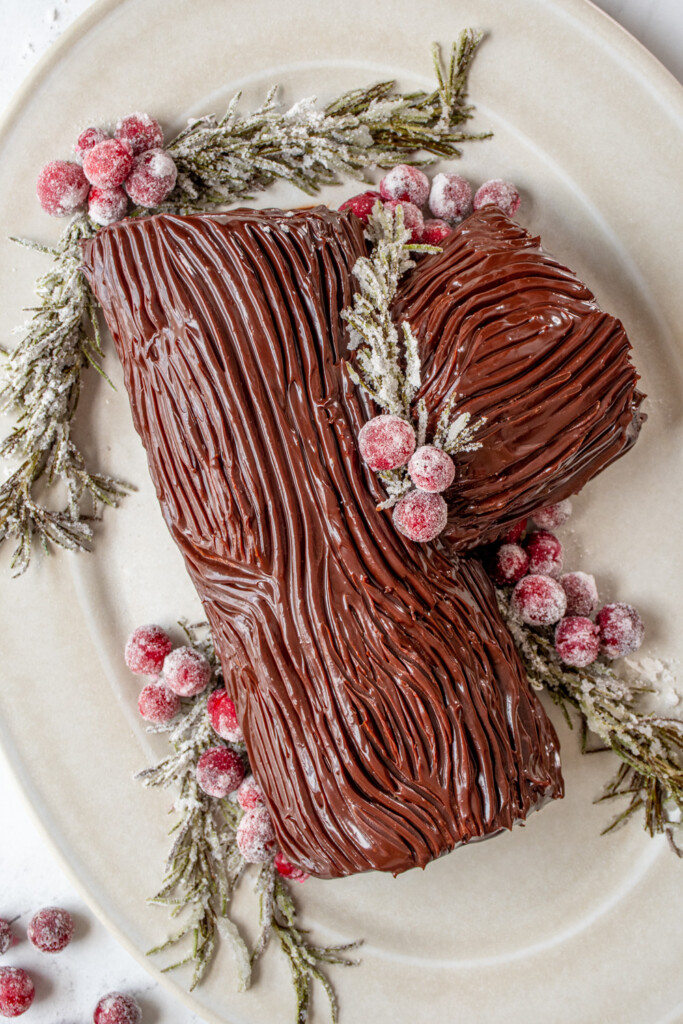 Overhead view of a yule log cake garnished with sugared cranberries and rosemary on a platter showing off the chocolate ganache that looks like tree bark.