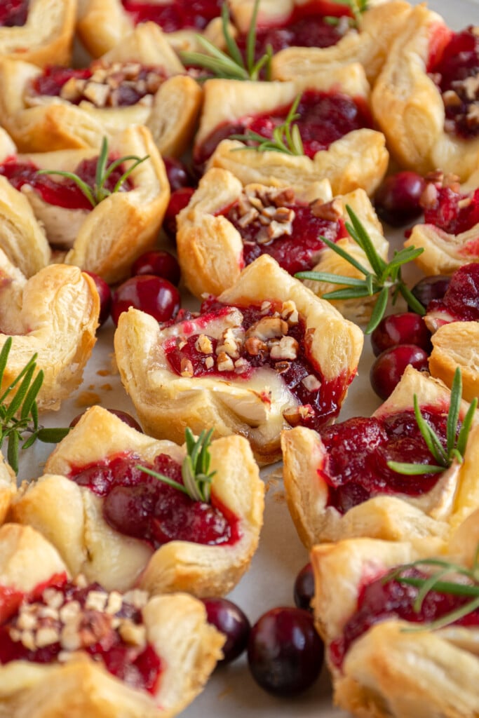 Close up of a cranberry brie bite on a platter surrounded by many other and springs or rosemary and fresh cranberries.