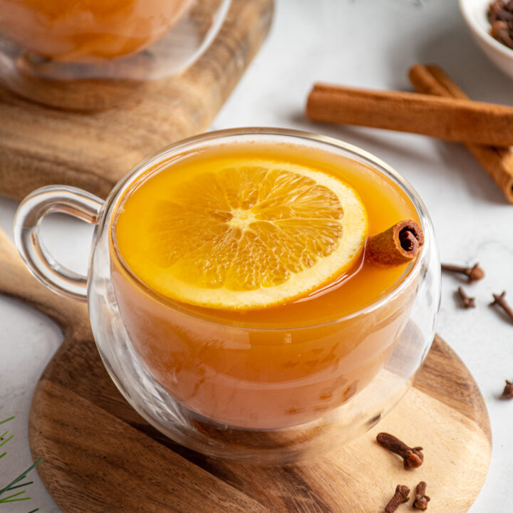 A glass mug filled with crockpot Christmas wassail with an orange slice floating on top next to a cinnamon stick.
