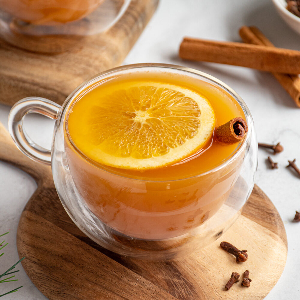 Clear mug filled with orange colored Christmas wassail with an orange slice and cinnamon stick as garnish.