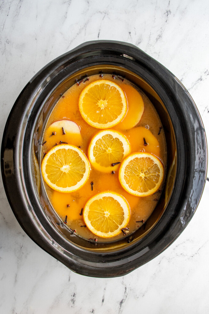A black crockpot with the garnishes in the wassail including apple & orange slices, cinnamon sticks, and cloves.
