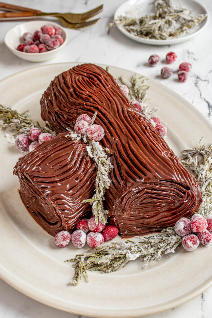 Yule Log Cake on a white platter garnished with sugared cranberries and rosemary.