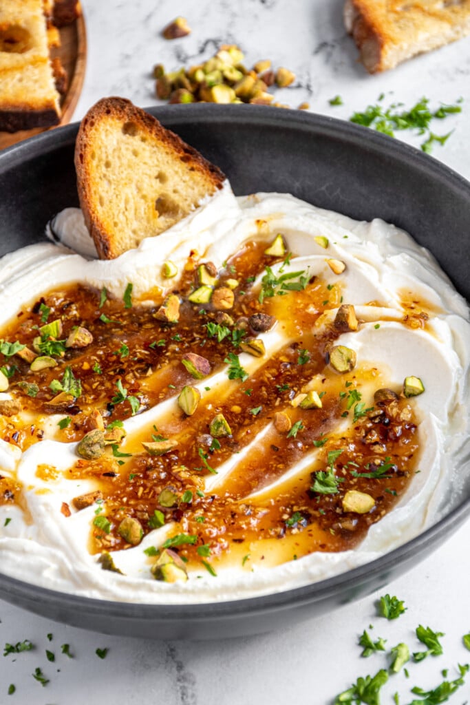 Bowl of whipped feta topped with honey and garnished with pistachios and parsley with a toast being dipped into the dip.