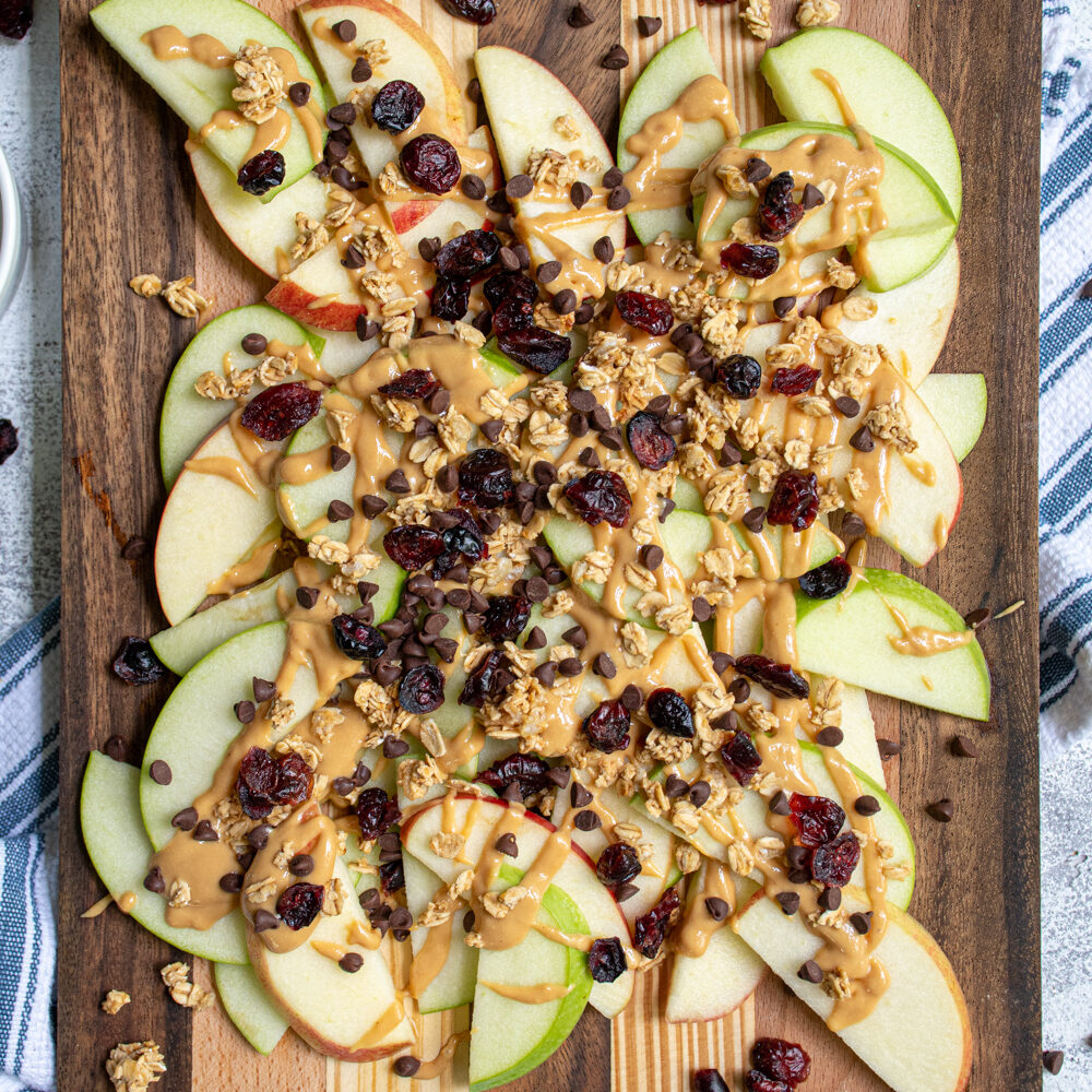 Overhead view of a wood cutting board piled high with apple slices covered in peanut butter, granola, dried cranberries, and mini chocolate chips.