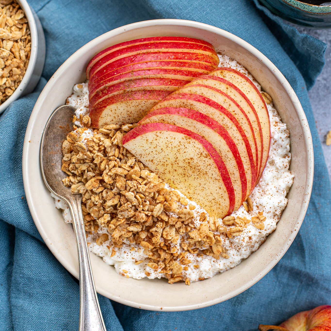 A white bowl on a blue towel filled with cottage cheese and apples with granola and ground cinnamon with a spoon ready to eat.