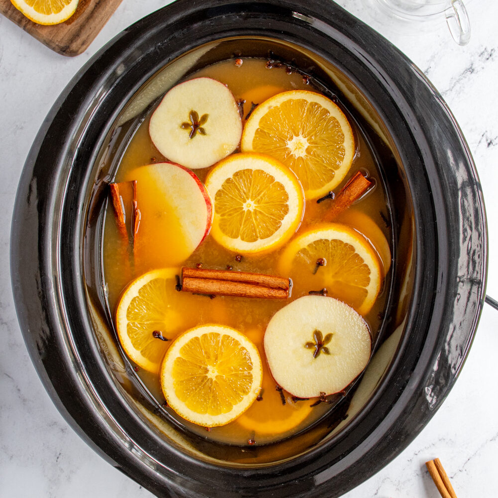 A black crockpot filled with Christmas wassail topped with apple and orange slices, cinnamon sticks, and cloves.