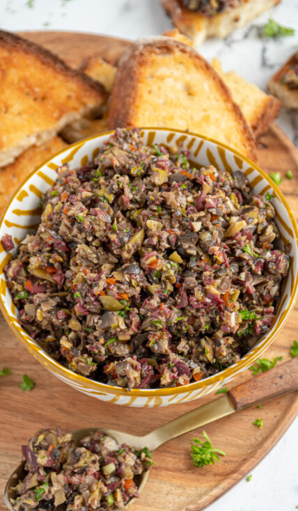 A yellow and white bowl filled with olive tapenade on a wooden serving board with pieces of toasts all around for serving.