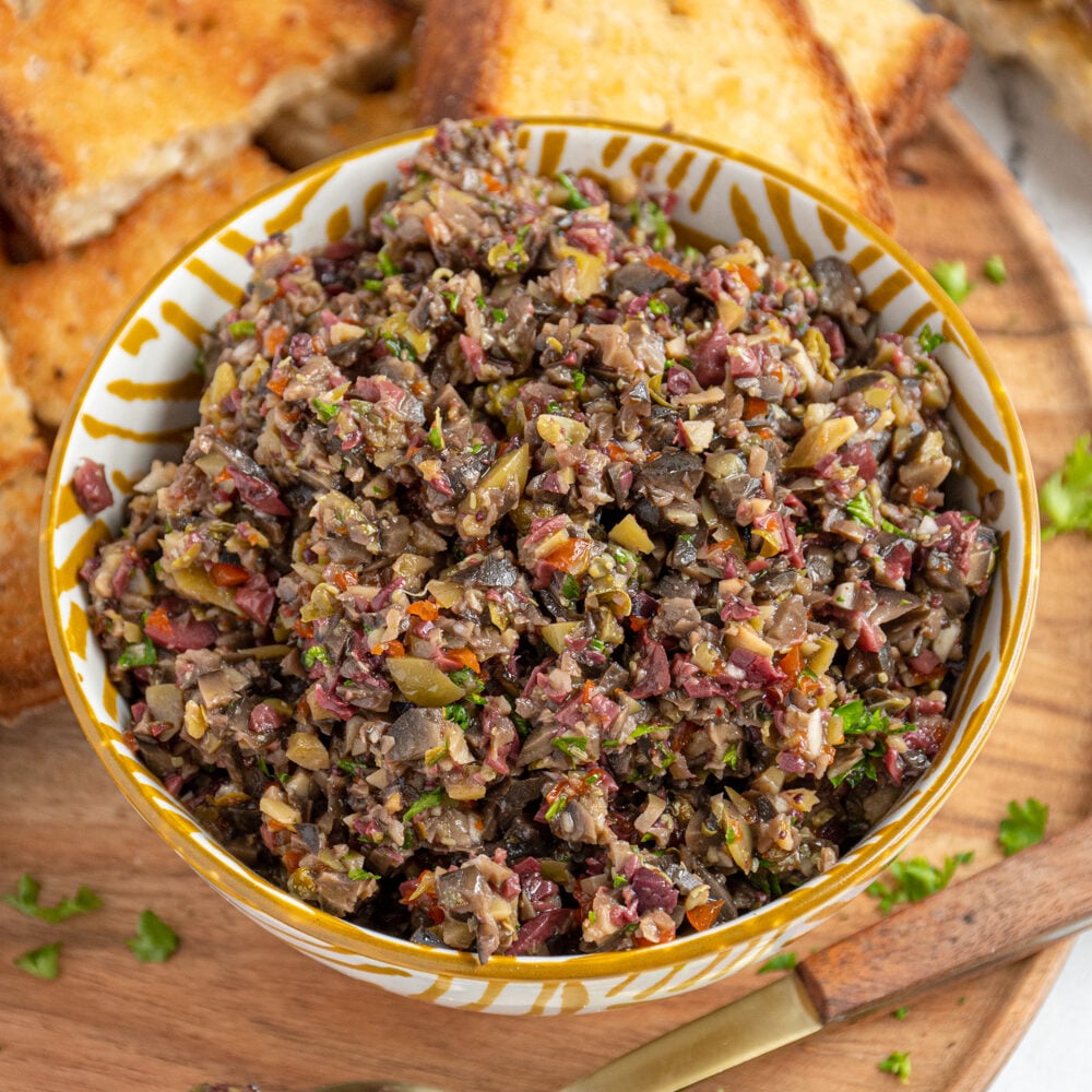 A yellow and white bowl filled with olive tapenade on a wooden serving board with pieces of toasts all around for serving.