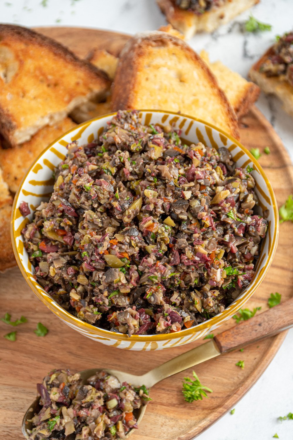 A yellow and white bowl filled with olive tapenade on a wooden serving board with pieces of toasts all around for serving.