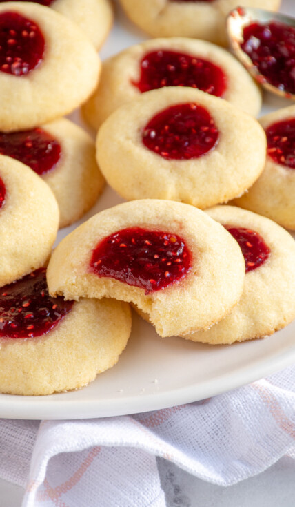 A plate of raspberry thumbprint cookies with the cookie the middle has a bite taken out revealing a soft dough.