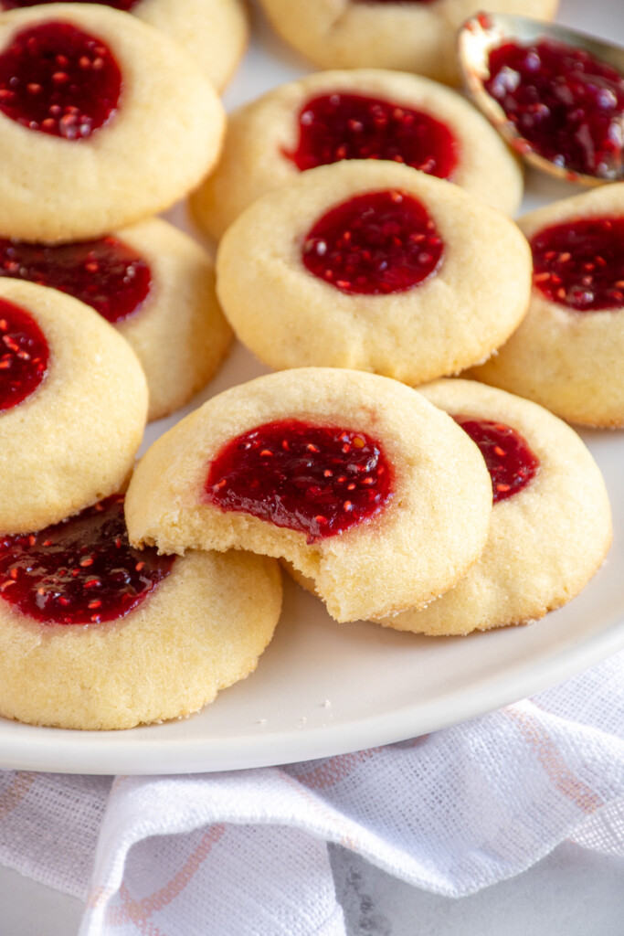 A plate of raspberry thumbprint cookies with the cookie the middle has a bite taken out revealing a soft dough.