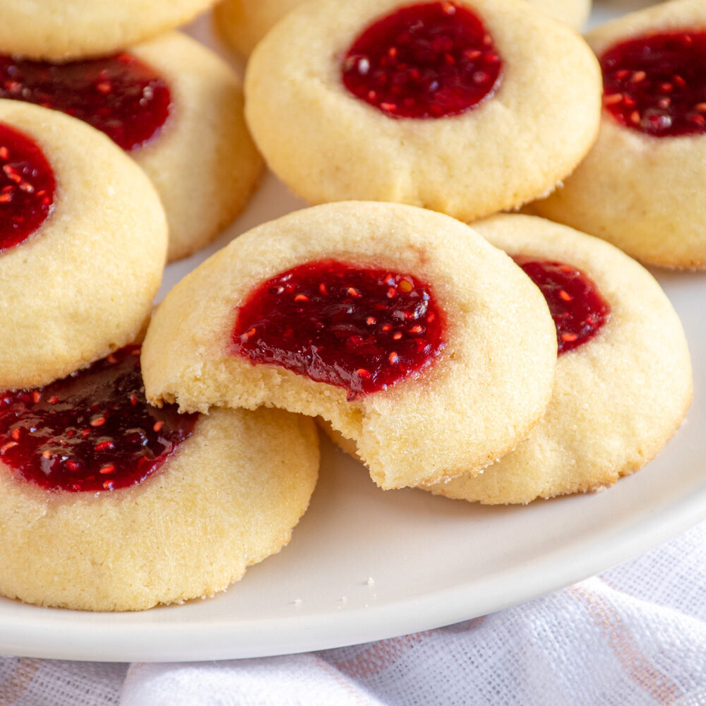 A plate of raspberry thumbprint cookies with the cookie the middle has a bite taken out revealing a soft dough.