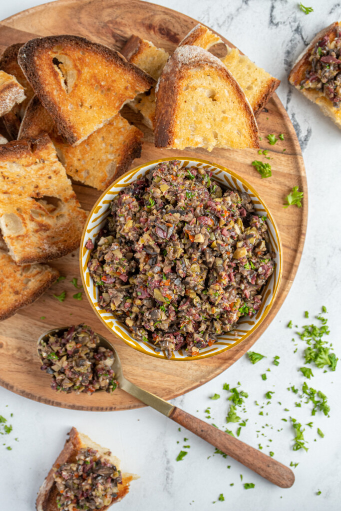Overhead view of a wooden serving platter with pieces of toast and a bowl filled with homemade olive tapenade.