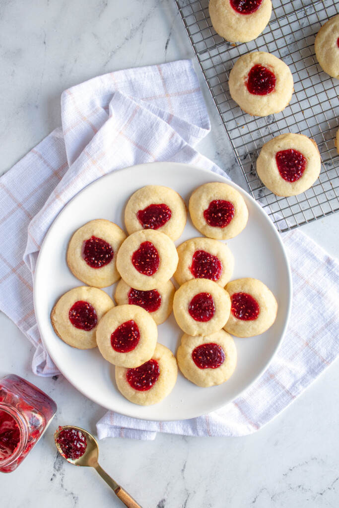 Overhead view of a white plate full of raspberry thumbprint cookies with bright red jam in the center.