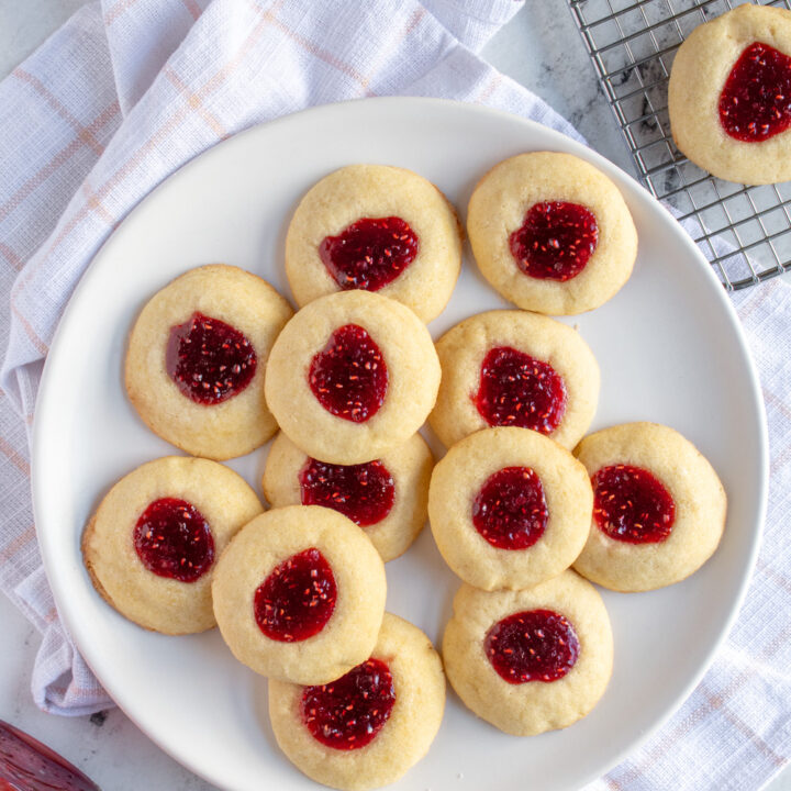 Overhead view of a plate of raspberry thumbprints with bright red jammy centers inside a soft sugar cookie.