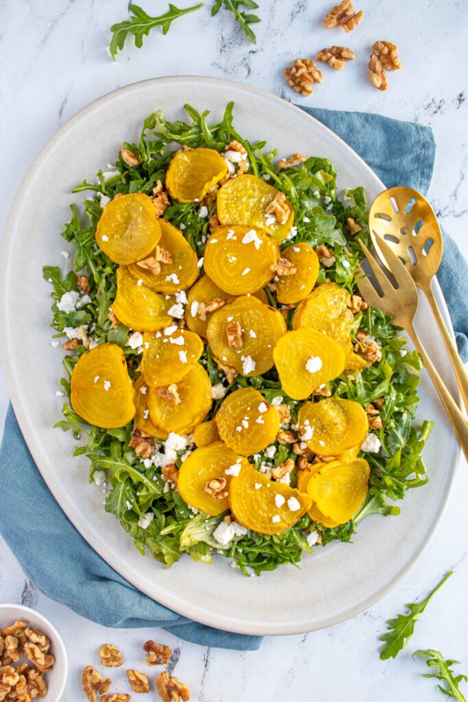 Overhead view of a white platter filled with arugula and topped with roasted golden beets, walnuts, and feta cheese.