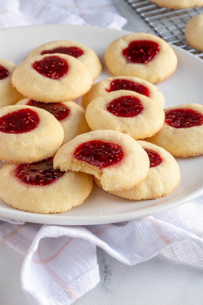 Platter of raspberry thumbprint cookies with red jam centers and soft sugar cookie bases, the one in the middle has bite taken out.